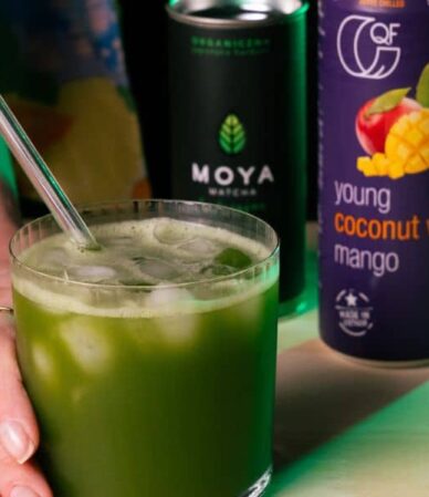 Mrożona matcha z lodem i słomką w szklance trzymanej w dłoniach, obok puszek z wodą kokosową QF Mango oraz Moya Matcha.